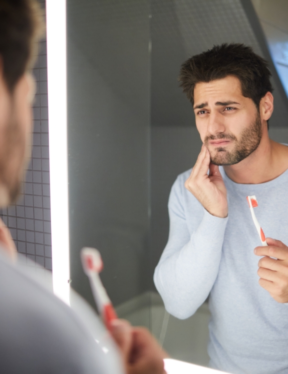 Man with toothbrush in bathroom holding his cheek in pain before emergency dentistry in Succasunna